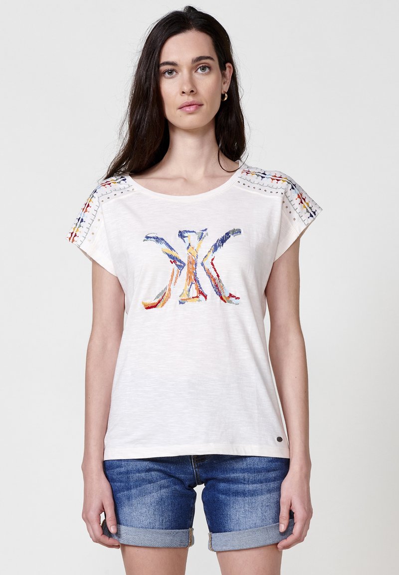 T-shirt court à manches courtes de couleur beige clair avec un design coloré brodé sur le devant et des accents à motifs sur les épaules ; fabriqué en tissu léger.