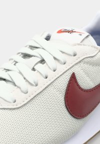 Svetlošedé tenisky s textúrovaným sieťovaným zvrškom, biele šnúrky a výrazný marhuľový swoosh logo po boku. Viditeľná gumová podrážka.