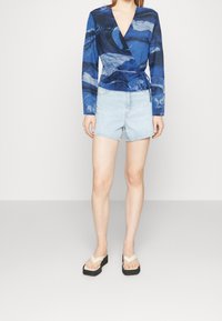 Top blu con maniche lunghe e stampa astratta a onde, design avvolgente, abbinato a pantaloni corti in jeans chiari e infradito nere con plateau.