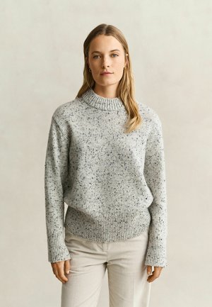 Vrouw met lang blond haar draagt een grijs gespikkelde gebreide trui en lichtbeige broek tegen een effen lichte achtergrond.
