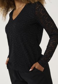 Top nero in pizzo a maniche lunghe con scollo a V, caratterizzato da motivi floreali e maniche trasparenti. Tessuto texturizzato e leggero.