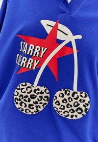 Modrá mikina s veľkou červenou hviezdou a nápisom "STARRY CHERRY", s plyšovými čerešňami v leopardom vzore ako významnými detailmi.