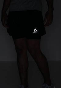 Černé sportovní šortky s reflexním logem Reebok, lehká tkanina, délka nad kolena, nošeny s šedými teniskami, na tmavém pozadí.