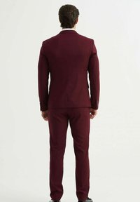 Costume bordeaux avec un design ajusté, présentant un revers à cran et un pantalon slim. Le tissu semble lisse, mettant en avant un look sur mesure.