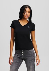 Tricou negru cu decolteu în V din material moale, cu mâneci scurte și un mic logo colorat pe pieptul stâng. Asociat cu blugi gri deschis.