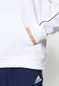 Sweat à capuche blanc avec une poche avant, texture douce, et bordure noire le long des manches, associé à un pantalon marine avec un logo Adidas.