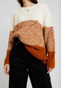 Gruby sweter robiony na drutach w odcieniach bieli, rdzawych i pomarańczowych w ombre. Ma szeroki dekolt i ściągacze. Teksturowana tkanina dodaje głębi.