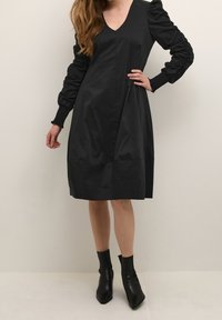 Vestido negro con escote en V y mangas abullonadas, con una textura suave y un diseño hasta la rodilla. Llevado con botas negras de tacón puntiagudas.