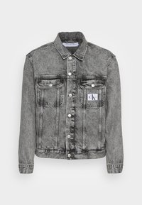 Grå denimjacka med knäppning fram, två bröstfickor och en Calvin Klein-logotyp, mot en enkel vit bakgrund.