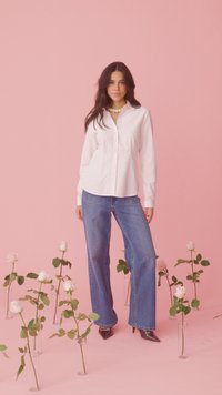 Femme aux longs cheveux noirs debout parmi des vases en verre contenant des roses blanches, portant une chemise blanche à rayures, un jean bleu et des talons noirs sur fond rose.