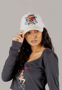 Weißer Hut mit buntem Totenkopf- und Herzgrafik; Netzrücken, gebogener Schirm; getragen mit einem langärmligen dunklen Oberteil mit Grafikdruck.