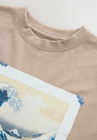 Camiseta de algodón beige con un gráfico cuadrado de olas rompiendo en azul y blanco, rodeado por un suave marco blanco.