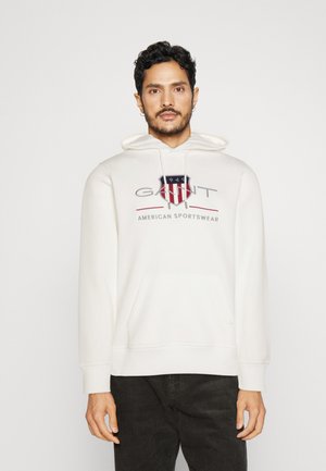 Mand iført hvid hættetrøje med GANT American Sportswear-logo, stående mod en ensfarvet lys baggrund, hænder afslappet ned langs siderne.