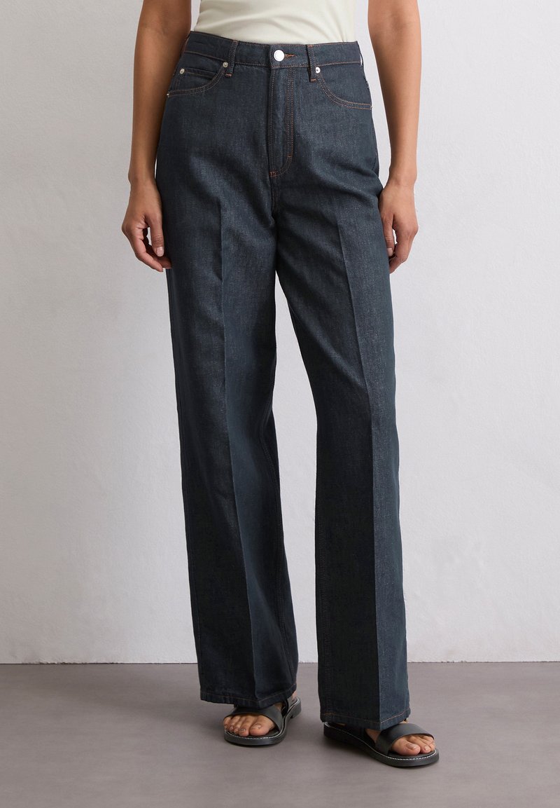 Ciemnoniebieskie szerokie spodnie dżinsowe z wysokim stanem, kontrastującym pomarańczowym przeszyciem i dwiema przednimi kieszeniami. Gładki materiał z delikatnym połyskiem.