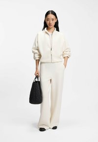 Weiße Reißverschlussjacke mit überschnittenen Schultern, kombiniert mit weiten weißen Hosen. Schwarze gesteppte Handtasche. Glatte Texturen und neutrale Farben.