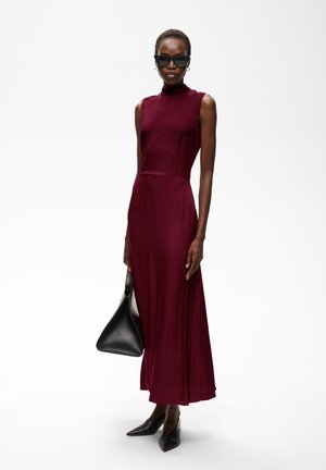 Femme portant une robe bordeaux sans manches, des chaussures noires à bout pointu, un sac à main noir et des lunettes de soleil foncées, debout sur fond blanc.