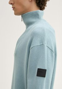Helles blauer Pullover mit hohem Rippkragen, Reißverschluss und strukturiertem Strickstoff. Schwarzes Etikett-Detail am Ärmel.