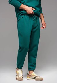 Pantalones de chándal en verde azulado con puños y cintura elásticos, que cuentan con un cordón. Combinados con zapatillas beiges y verdes con acentos texturizados.