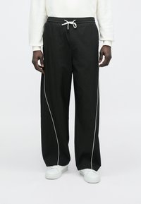 Μαύρο, φαρδύ παντελόνι με κορδόνι στη μέση και λευκή ρίγα στο πλάι, κατασκευασμένο από ελαφρύ υλικό, σε συνδυασμό με λευκά αθλητικά παπούτσια.