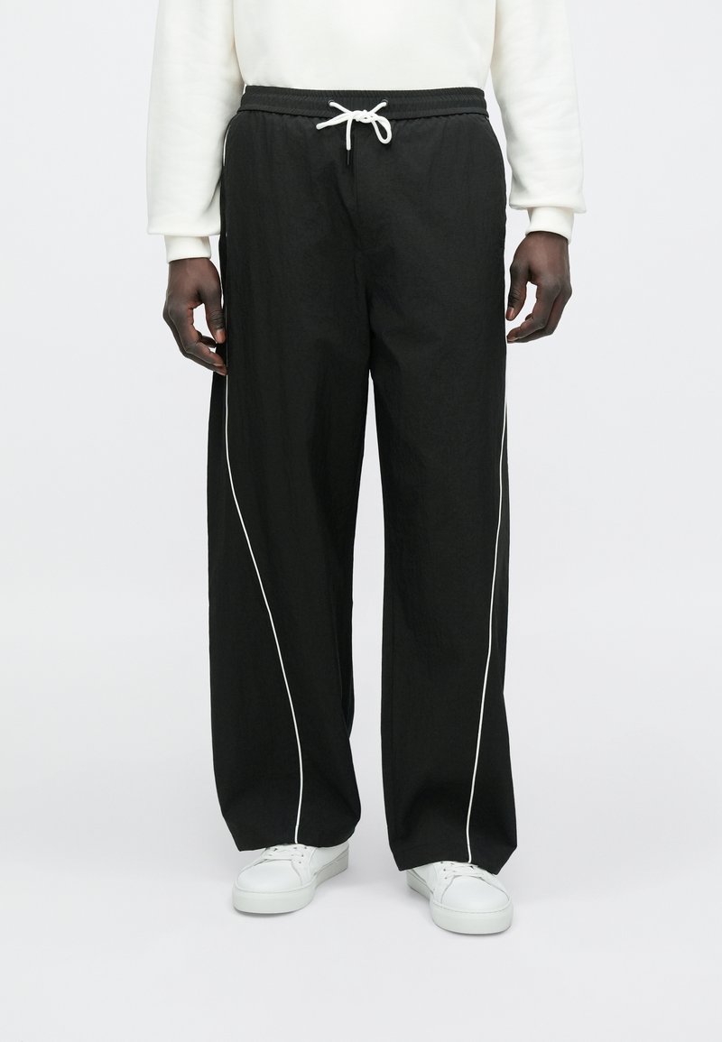 Μαύρο, φαρδύ παντελόνι με κορδόνι στη μέση και λευκή ρίγα στο πλάι, κατασκευασμένο από ελαφρύ υλικό, σε συνδυασμό με λευκά αθλητικά παπούτσια.