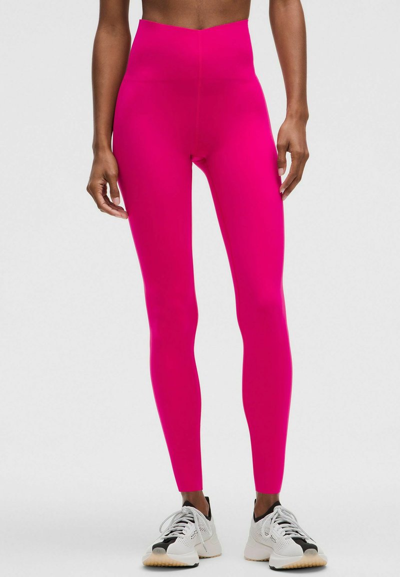 Magasan fazonú, élénk rózsaszín leggings, rugalmas anyagból, sima felülettel. Fehér és fekete sportcipővel viselve.