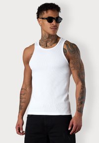 White tank top made of ribbed cotton, featuring a round neckline and fitted design, worn with black sunglasses, showing arm tattoos.