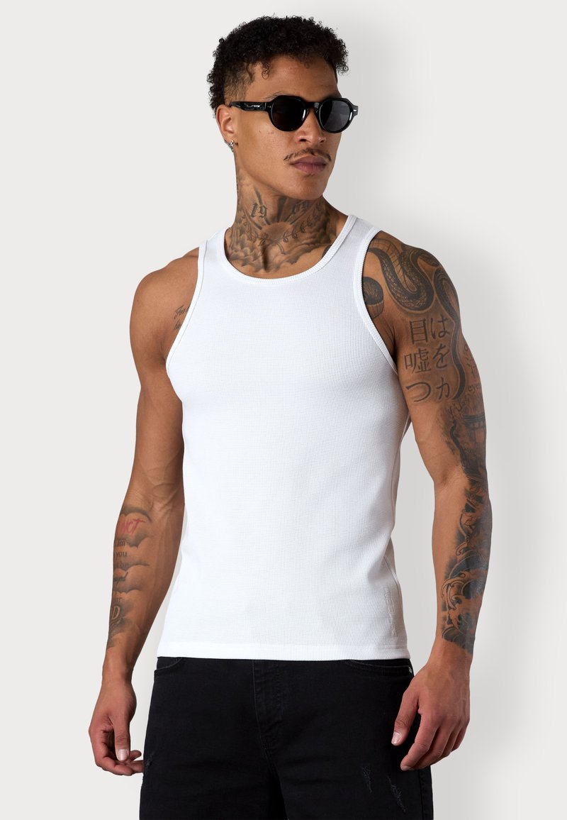 White tank top made of ribbed cotton, featuring a round neckline and fitted design, worn with black sunglasses, showing arm tattoos.