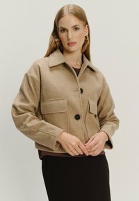 Giacca di lana corta color sabbia con chiusura a bottoni, due tasche sul petto e colletto; presenta una texture liscia e un design minimalista.