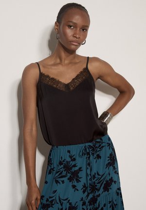 Mujer con camisola negra con encaje y falda azul con estampado floral negro, posando con la mano en la cadera frente a un fondo liso.