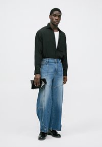 Chemise noire à manches longues, t-shirt blanc en dessous, jeans larges en denim bleu, tenant une pochette noire, bottes noires, tenue décontractée.