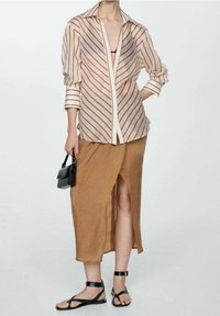 Camicia a maniche lunghe a righe in tessuto leggero con righe diagonali rosse e bianche, abbinata a una gonna marrone con spacco fino alla coscia e sandali neri.