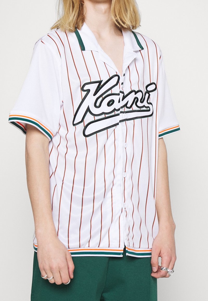 Chemise blanche à manches courtes avec des rayures verticales rouges et des accents verts. Affiche le nom de la marque "Kani" en script noir en gras.