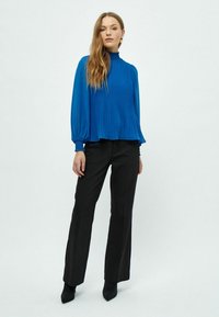 Blusa plissettata blu con collo alto e maniche lunghe, abbinata a pantaloni neri su misura, caratterizzati da una texture liscia e una silhouette aderente.