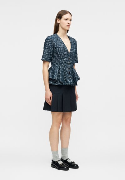 Femme portant un haut péplum texturé bleu marine, une jupe plissée noire, des chaussettes grises et des chaussures noires à boucle, debout devant un fond uni.