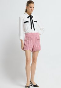 Bílá halenka s černými akcenty a mašlí, v kombinaci s růžovými vzorovanými šortkami s dvěma předními kapsami a zlatými knoflíky. Na nohou jsou černé boty, které outfit doplňují.
