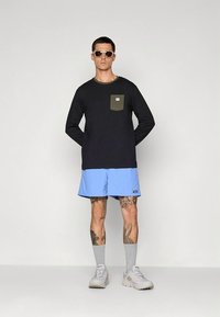 Camisola preta de manga comprida com bolso verde-oliva, combinada com calções azul claro. O modelo usa ténis brancos e meias cinzentas, com óculos de sol.