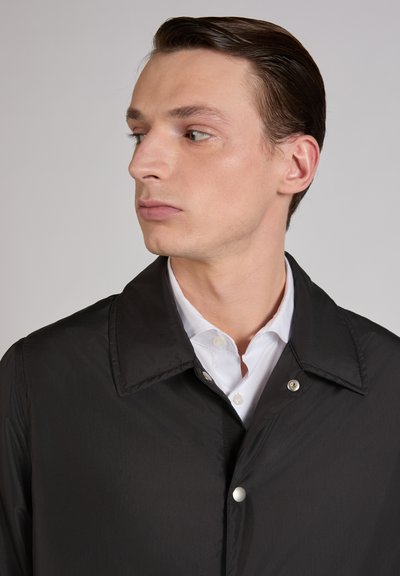 Młody mężczyzna z zaczesanymi do tyłu włosami, ubrany w czarną kurtkę na białej koszuli zapinanej na guziki, patrzący w lewo na tle jednolitego tła.