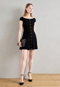 Robe noire à côtes avec de courtes manches dénudées, ornée de boutons dorés sur le devant et d'une jupe évasée. L'accessoire est une pochette noire.