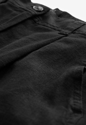 Gros plan sur un pantalon en tissu noir montrant le bouton avant, la ceinture et une poche latérale partielle avec des coutures et une texture visibles.