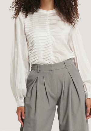 Woman wearing a white long-sleeve blouse with horizontal pleats and gray high-waisted pleated trousers.