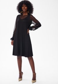 Frau mit lockigem Haar, die ein knielanges schwarzes Kleid mit durchsichtigen langen Ärmeln und beigen High Heels trägt, steht vor einem weißen Hintergrund.