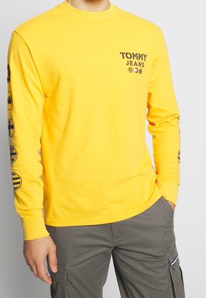 Mann trägt ein gelbes Langarmshirt mit dem Schriftzug "Tommy Jeans" auf der Brust und mehreren runden Flaggenmotiven auf den Ärmeln, kombiniert mit grauen Cargo-Hosen.