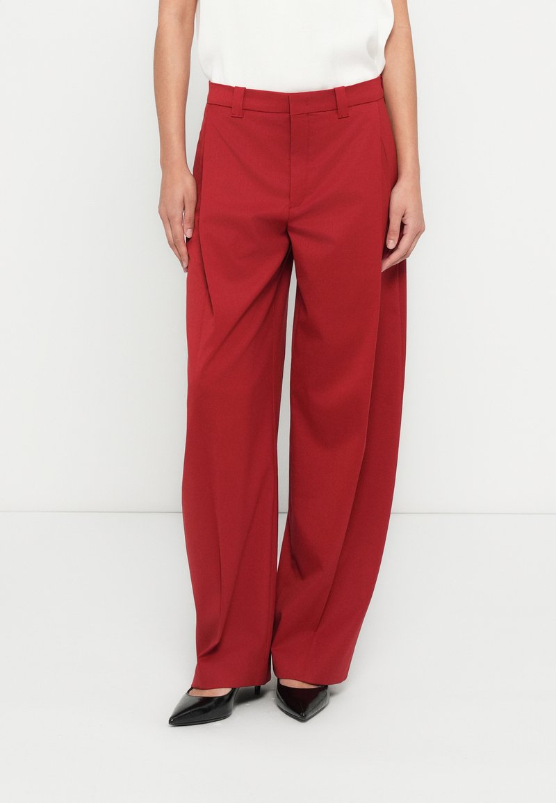 Pantalon rouge à jambes larges en tissu lisse, coupe ajustée, taille haute et poches latérales, porté avec des escarpins noirs.