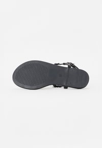 Svart sandal med teksturert såle, med tynne stropper og metallakenter. Designet er flatt, som gir åpne tær fleksibilitet.