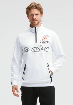 Man with light brown hair wears white zipped sweatshirt with black zippers and "Gaastra" logo, paired with black pants, standing against plain background.