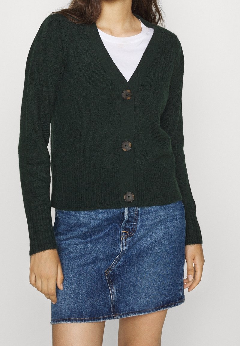 Femme portant un cardigan boutonné vert foncé sur un haut blanc assorti d'une mini-jupe en jean bleu, debout les mains détendues sur les côtés.