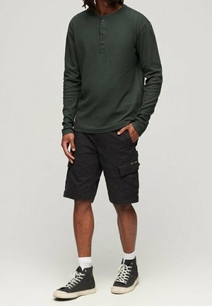 Mężczyzna ubrany w ciemnozieloną koszulę henley z długim rękawem, czarne szorty z kieszeniami, białe skarpetki i czarne wysokie trampki, stoi z jedną ręką w kieszeni.