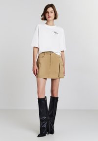 T-shirt blanc en coton avec logo, associé à une jupe en denim beige avec poches cargo, complété par des bottes en cuir noir montantes.