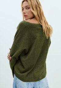 Jonge vrouw met lang blond haar, gekleed in een losse groene trui met blote schouders en een lichtblauwe denim rok, kijkt over haar schouder.