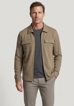 Man met een beige jas met rits en klepzakken, donkergrijs overhemd en beige broek, staand tegen een effen lichte achtergrond.